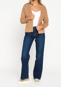 Cardigan beige de felpa con apertura frontal, llevado sobre una blusa blanca. Combinado con jeans oscuros de cintura alta y zapatillas de color claro.
