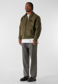 Olivgrön bomberjacka med dragkedja framtill, ribbade ärmslut och broderad logga. Buren över en vit T-shirt och parat med grå lösa jeans och svarta skor.