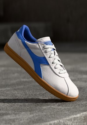 Baskets blanches et bleues avec semelle en gomme, suspendues au-dessus d'une surface grise texturée avec une ombre en dessous.