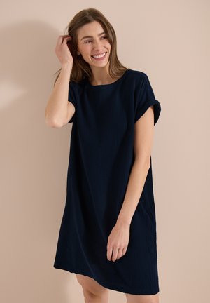 Jeune femme souriante, portant une robe noire ample à manches courtes, debout devant un fond beige uni.