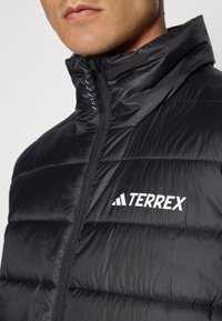Black puffer jacket with a high collar, quilted texture, zip closure, and white "TERREX" logo on the chest. Lightweight material.
