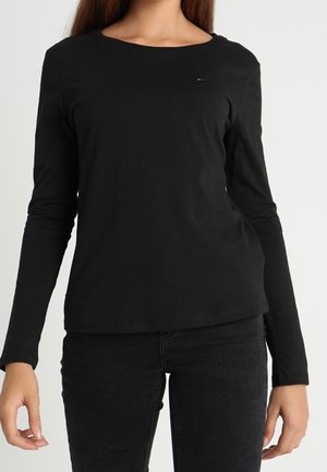 Frau trägt ein eng anliegendes schwarzes Langarmshirt und schwarze Hose, steht vor einem schlichten hellen Hintergrund.