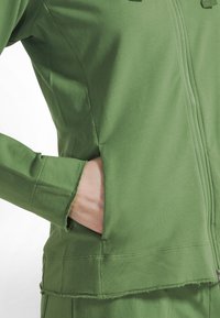 Chaqueta verde con cremallera confeccionada en tela suave, con bolsillo frontal, mangas con puño y detalles de dobladillo sin rematar para un look relajado.