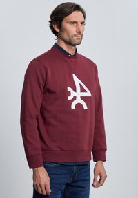 Sudadera burdeos con un logotipo abstracto blanco, cuello redondo y puños acanalados. La tela tiene una textura suave. Llevada sobre una camisa azul con cuello.