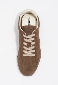 Brązowe zamszowe sneakersy z kremowymi sznurówkami, falisty wzór wzdłuż krawędzi oraz teksturowana podeszwa. W środku znajdują się nadrukowane logo.