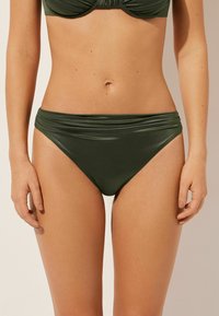 Zelené bikiny s hladkým, elastickým materiálem. Mají široký pás s nařasenými detaily. Středně vysoký a těsně přiléhající střih.