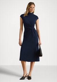 Srednje dolga mornarsko modra obleka z visokim izrezom, kratkimi rokavi in naborki na pasu. Model drži črno torbico.