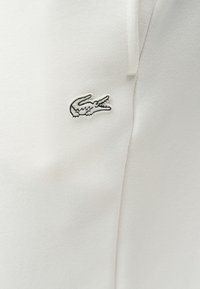 Λευκό ύφασμα με μικρό κεντημένο μαύρο και λευκό λογότυπο κροκοδείλου στην αριστερή πλευρά, που υποδεικνύει μια λεπτομέρεια ρούχου επωνυμίας.