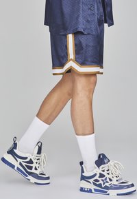 Marinefarbene Sportshorts mit weißen und goldenen Streifen, kombiniert mit marineblauen und weißen Sneakers sowie weißen Socken. Kurzarmshirt aus strukturiertem Material.