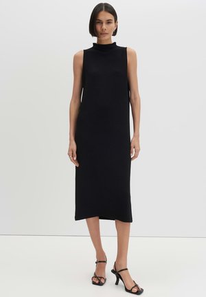 Femme aux cheveux courts et foncés, portant une robe midi noire sans manches et des sandales à talons hauts noires à brides, debout sur un fond neutre.