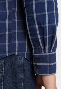 Chemise bleu marine à boutons avec un motif écossais, comportant des lignes bleu clair contrastées, un tissu texturé, et un détail de poignet à double bouton.