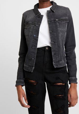 Personne portant une veste en jean noire délavée par-dessus une chemise blanche avec un jean noir déchiré sur un fond gris uni.