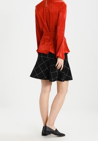 Top lungo rosso in satin con maniche a sbuffo e orlo peplum abbinato a una gonna nera con motivo a griglia bianca e ballerine nere.