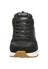 Sneaker nera con una combinazione di materiale scamosciato e pelle, caratterizzata da un design con lacci neri, suola intermedia bianca e suola in gomma color caramello.