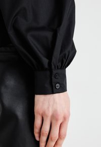 Polsino di camicia nera con tessuto arricciato, due bottoni neri e una superficie liscia. Mano appoggiata su una superficie nera simile alla pelle.