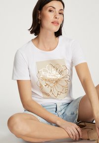 Biała, krótka koszulka z krótkim rękawem z złotym wzorem kwiatowym na beżowym kwadracie, w połączeniu z jasnoniebieskimi szortami dżinsowymi i brązowym obuwiem.