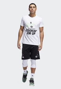 Camiseta blanca con texto negro "COOKIN' EM UP" y gráfico, combinada con pantalones cortos negros. El modelo lleva una malla blanca de compresión y zapatillas estampadas.