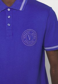 Fialová polo košile s žebrovaným límečkem, zapínáním na knoflíky a kruhovým vyšitým logem s nápisem "Versace Jeans Couture." Kontrastní pruhy na límečku.