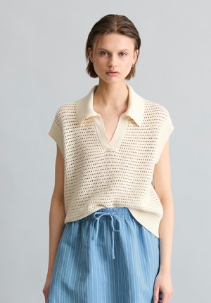 Jonge vrouw draagt een mouwloos crèmekleurig gebreid topje met kraag en een blauwe broek met fijne strepen en trekkoord, staand tegen een neutrale achtergrond.