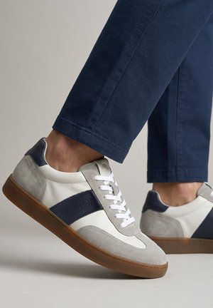 Füße in marineblauen Hosen und weißen Turnschuhen mit grauem Wildleder, blauem Streifen, weißen Schnürsenkeln und gummierten Sohlen, ein Fuß leicht angehoben.