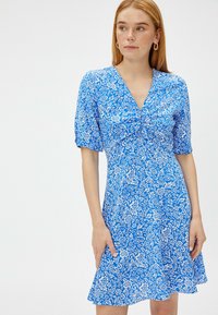 Blauwe jurk met bloemenprint, een V-hals en korte mouwen. De stof heeft een lichte textuur en een uitlopende onderkant met een speels ontwerp.