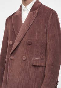 Homme portant un blazer en velours côtelé rose poussière à double boutonnage avec des boutons recouverts de tissu et une chemise blanche à col en dessous.