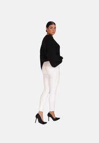 Pull en tricot noir, coupe ample, associé à un jean slim blanc. Le mannequin porte des chaussures à talons hauts noires. Couleurs unies, sans motifs.
