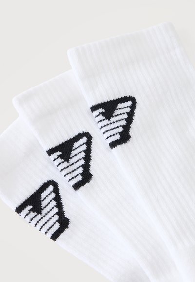 Trois chaussettes blanches côtelées avec un logo "V" rayé noir et blanc près du bord supérieur, posées en chevauchement sur un fond clair.