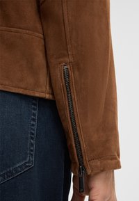 Bruine suède jas met een gestructureerde afwerking, met een ritssluiting bij de manchetten en een strakke uitstraling. Gecombineerd met donkere denim jeans.