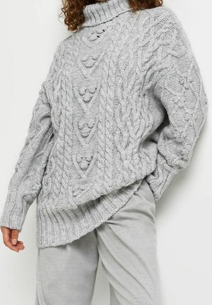 Femme portant un pull à col roulé en grosse maille torsadée gris clair et un pantalon assorti gris clair, posant avec une main dans la poche.