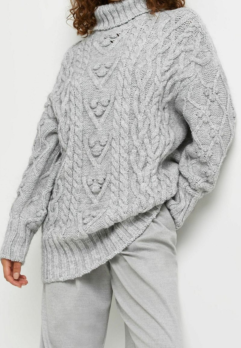 Femme portant un pull à col roulé en grosse maille torsadée gris clair et un pantalon assorti gris clair, posant avec une main dans la poche.