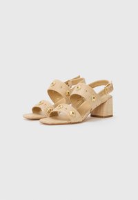 Beige block-heeled sandals made from textured material, featuring floral accents on straps. Adjustable ankle buckle for fit.