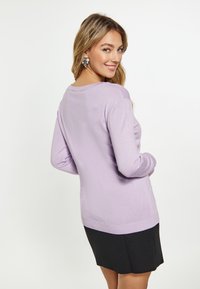 Maglione lungo con maniche a sbuffo di un delicato color lavanda, con polsini e orlo a coste. Indossato sopra una gonna nera. Tessitura liscia e vestibilità morbida. Orecchini d'argento completano l'outfit.