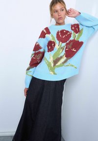Jeune femme portant un pull bleu clair orné de grands motifs de roses rouges et une longue jupe noire, posant contre un mur blanc.