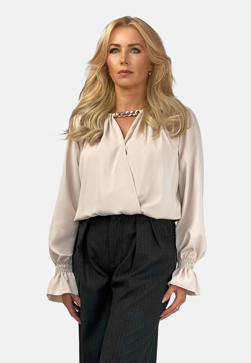 Jasnoróżowa bluzka z dekoltem w serek i detalem w postaci łańcuszka, z długimi rękawami i marszczonymi mankietami. Połączona z spodniami o wysokim stanie w ciemne paski.