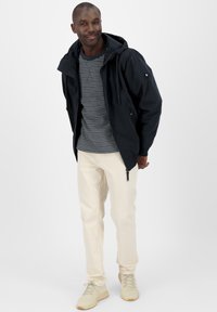 Giacca nera con cappuccio e lacci, maglietta a righe grigie e bianche, pantaloni color crema e sneakers beige chiaro. Outfit casual moderno.