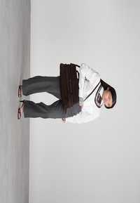 Γυναίκα με λευκό πουκάμισο και γκρι παντελόνι σε γραμμή, κρατώντας μια μεγάλη καφέ τσάντα, να στέκεται μπροστά σε έναν απλό γκρίζο τοίχο.