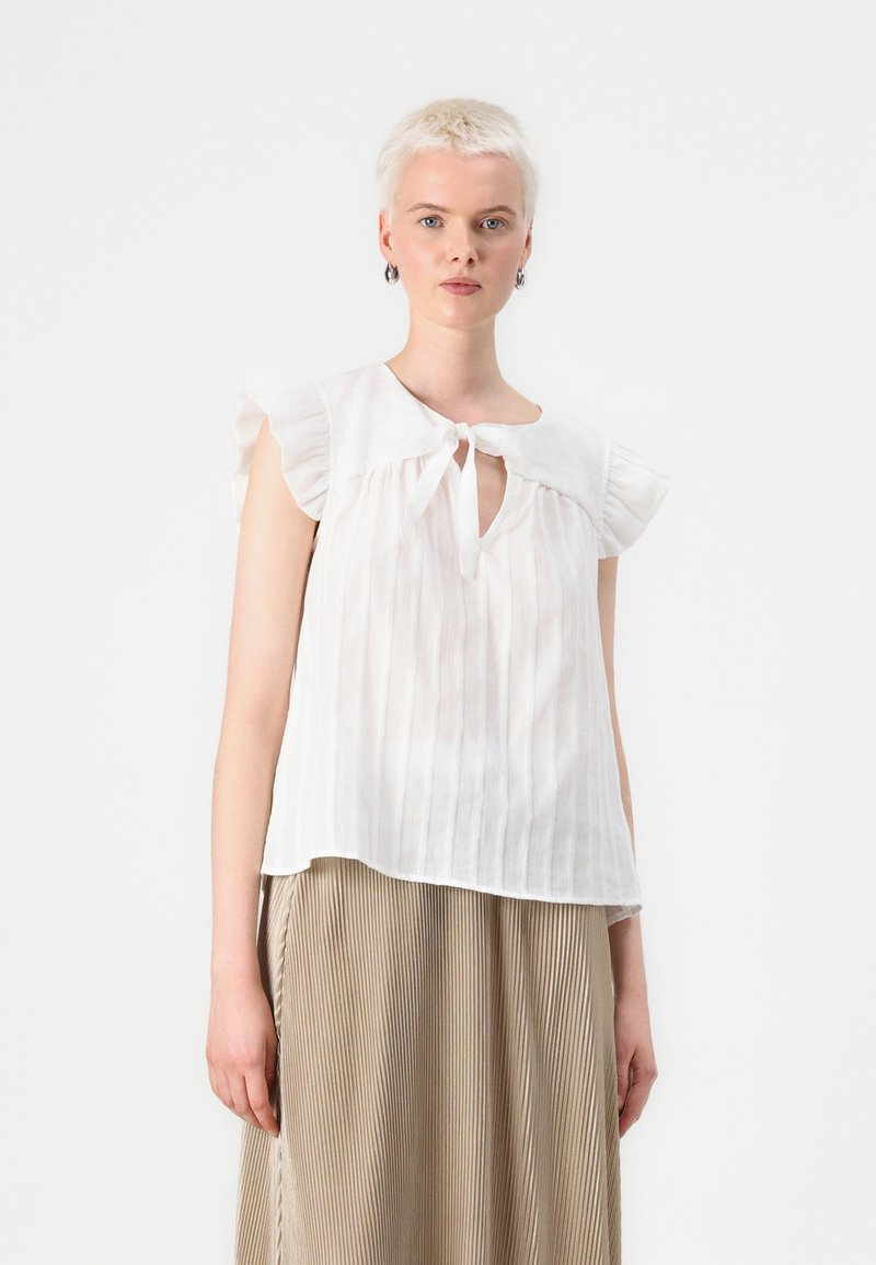 Blusa bianca leggera con dettaglio a chiave con nastro, maniche corte arricciate e righe verticali, abbinata a una gonna plissettata beige.