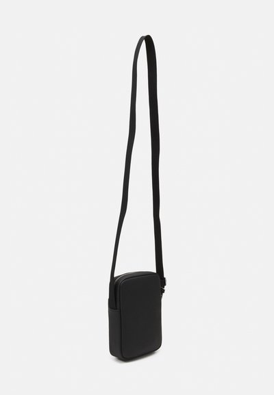 Sacoche noire à porter en bandoulière, de forme rectangulaire, avec une texture lisse, une fermeture éclair et une sangle ajustable. Design minimaliste sans motifs.