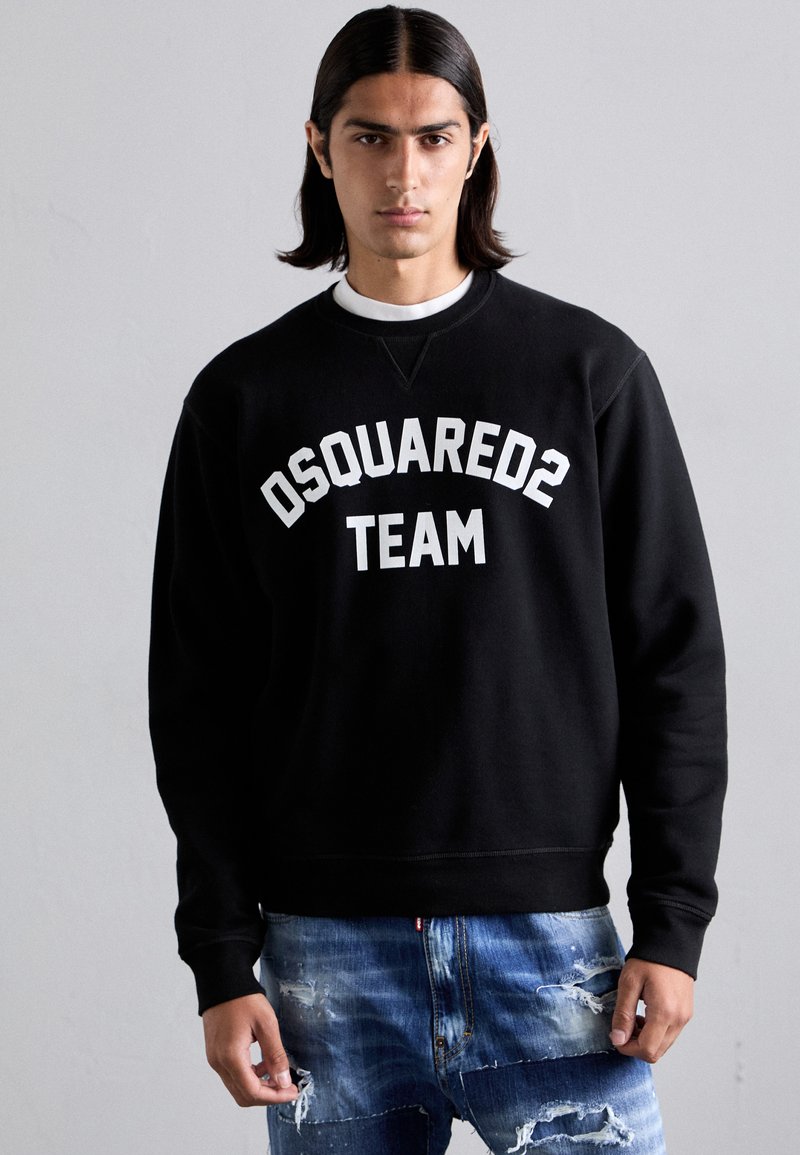 Hanorac negru cu inscripția "DSQUARED2 TEAM" imprimată în alb. Prezintă un guler rotund și manșete striate. Asortat cu blugi din denim albastru deteriorați.