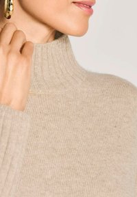 Nahaufnahme einer Person, die einen beigen gerippten Rollkragenpullover und einen goldenen Creolenohrring trägt und mit der Hand den Kragen berührt.