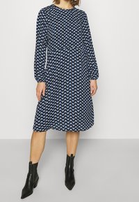 Långärmad blå klänning med vitt bladmönster, rund halsringning och en flödande silhuett, matchad med svarta ankelboots.