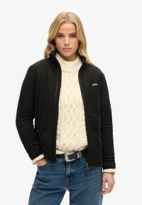 Veste en polaire noire avec zip sur le devant et col haut, portée sur un pull à torsades crème, associée à un jean en denim bleu.