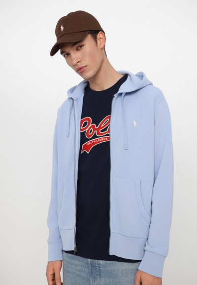 Sweatshirt azul claro com fecho zip, com um bolso canguru, sobre uma T-shirt azul marinho com um logótipo "Polo" bordado em vermelho. Boné castanho acima.
