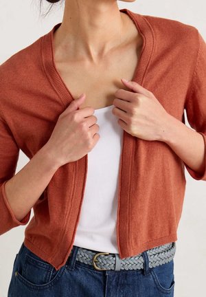 Femme ajustant un cardigan couleur rouille sur un haut blanc, portant une ceinture tressée grise et un jean bleu, vue des épaules à la taille.