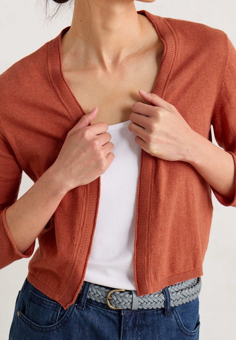 Femme ajustant un cardigan couleur rouille sur un haut blanc, portant une ceinture tressée grise et un jean bleu, vue des épaules à la taille.