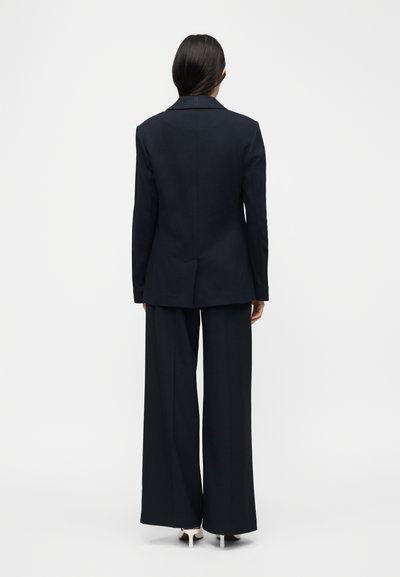 Γυναίκα με σκούρα μαλλιά φοράει μαύρο σακάκι προσαρμοσμένης κοπής και μαύρο παντελόνι με φαρδιές παντελόνες, στέκεται γυρισμένη προς τα πίσω σε απλό λευκό φόντο.