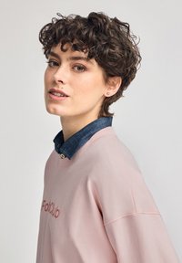 Felpa rosa con scollo rotondo, caratterizzata da un logo ricamato a trama. Sotto, è visibile una camicia collare in denim scuro.