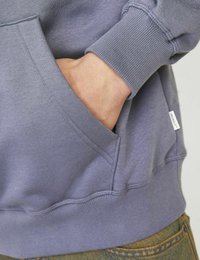 Lila pulcsi bordázott mandzsettákkal és aljjal, elöl zsebes kivitelben. Puha anyag és laza dizájn, részben látható kéz.