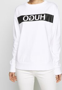 Bílá mikina s černým logem "HUGO" přes přední část. Má kulatý výstřih, žebrované manžety a lem. Měkká textura, pohodlný design.